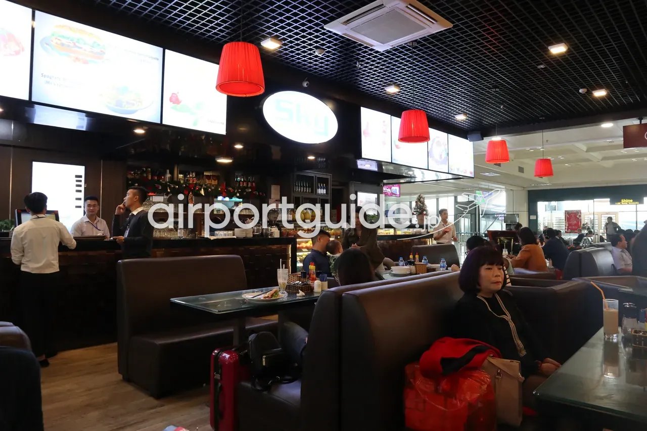 ノイバイ空港の「Sky」カフェ内部。緑に光る「Sky」のサイン、赤いペンダント照明、革張りのソファ席、クリスマスリースを飾ったコーヒー／ペストリーのカウンター、休憩する旅客。