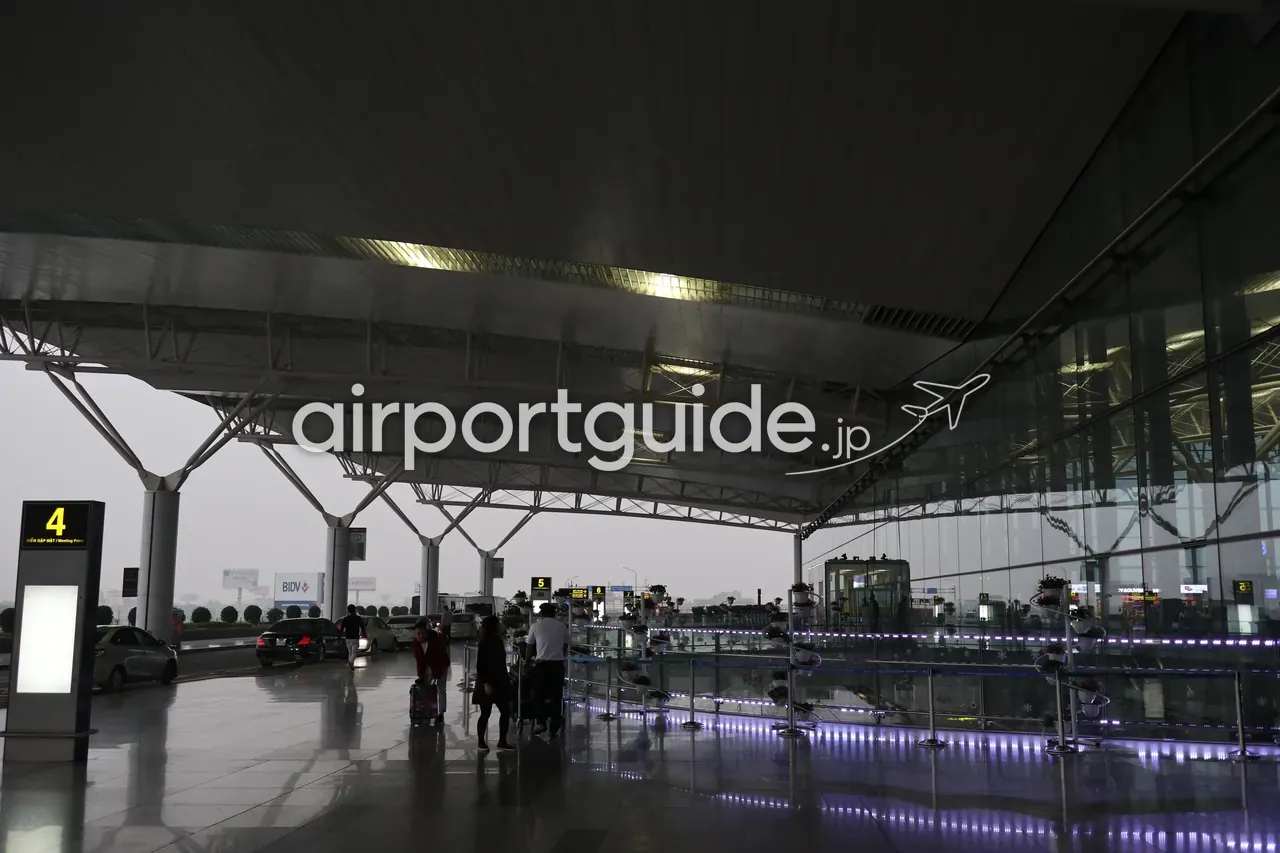 ノイバイ空港・第2ターミナル（国際線）出発階前の屋根付きフロント。霧がかった夕暮れに「4 ĐIỂM GẶP MẶT / Meeting Point（4番待ち合わせ場所）」の案内塔と「5」の柱、BIDVの広告、紫のLEDの手すり、旅客と車が写る。
