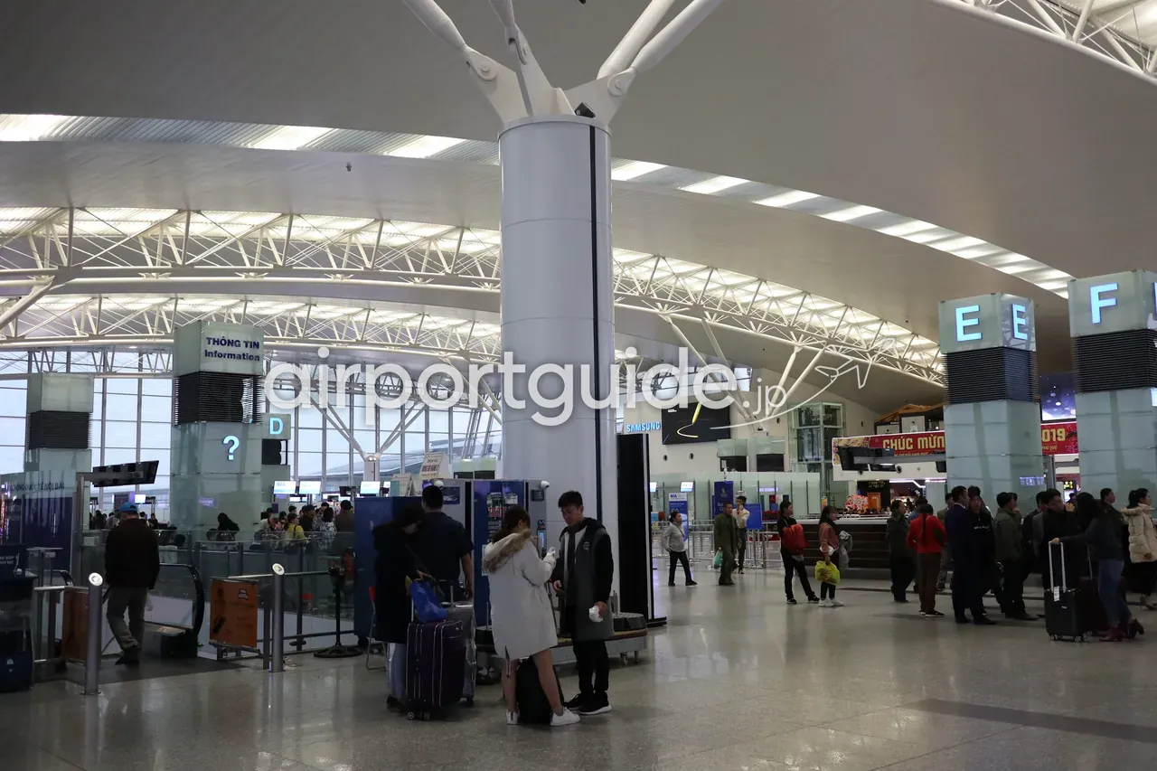 ノイバイ国際空港・第2ターミナル(国際線)の出発ホール。曲面トラスの大屋根の下、「THÔNG TIN / Information」の案内塔とゾーン表示「D」「E E F」、荷物カートを押す旅客が行き交う開放的な空間。