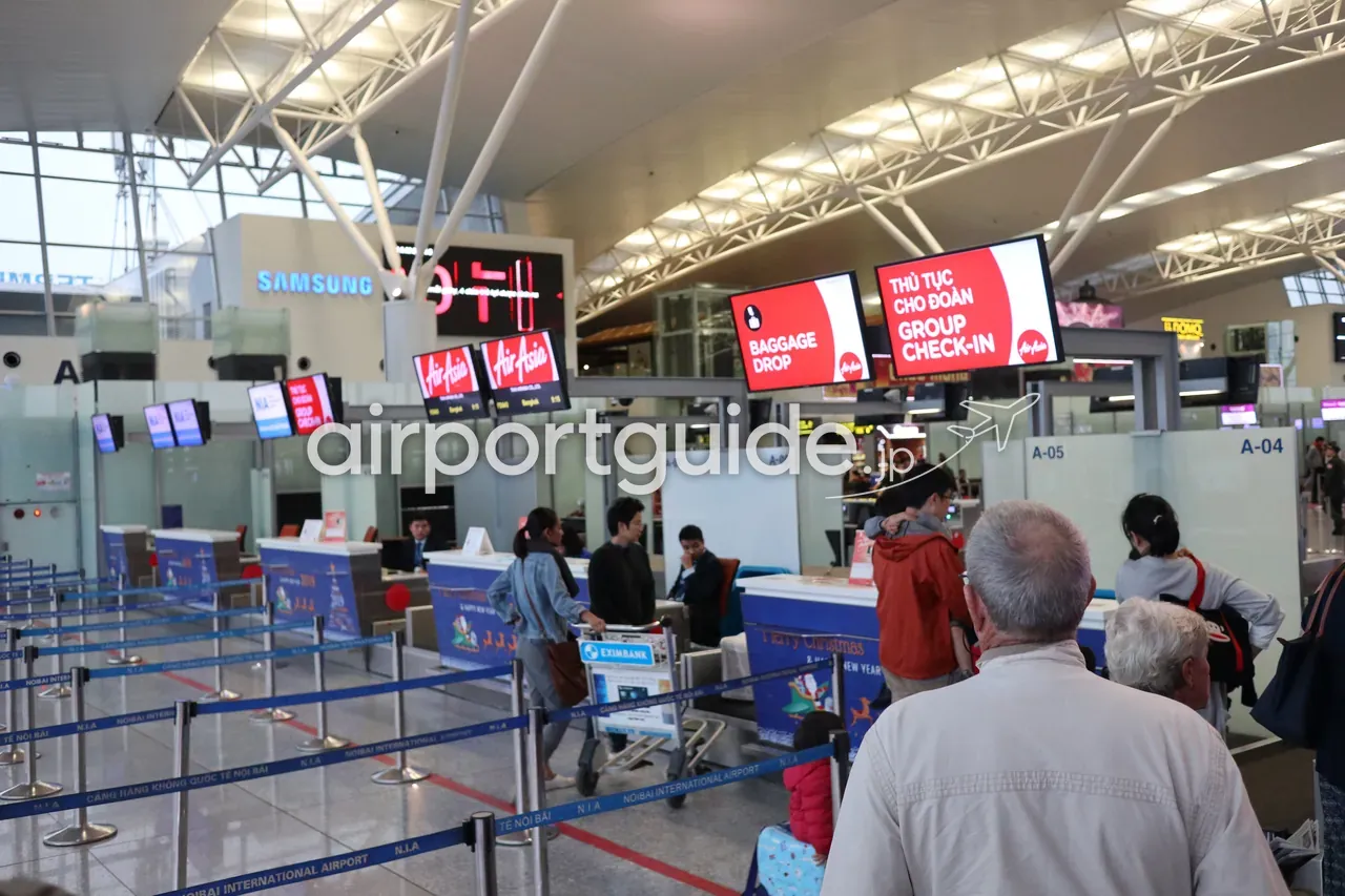 ノイバイ空港・第2ターミナル（国際線）のAirAsiaチェックインカウンター（A-04〜A-07付近）。「BAGGAGE DROP」「THỦ TỤC CHO ĐOÀN / GROUP CHECK-IN」の赤いサイン、曲面トラスの大屋根、N.I.Aの規制ベルト。