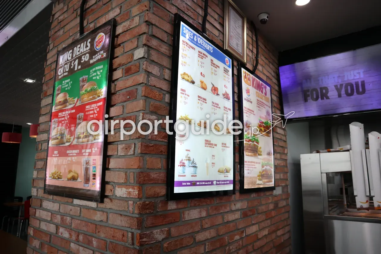 ノイバイ空港内バーガーキングのメニューボード。レンガ張りの柱に「KING DEALS（$1.50〜）」「SIDES & DESSERTS」「HOME OF THE WHOPPER」「ALL THAT, JUST FOR YOU」、価格はUSD表示。