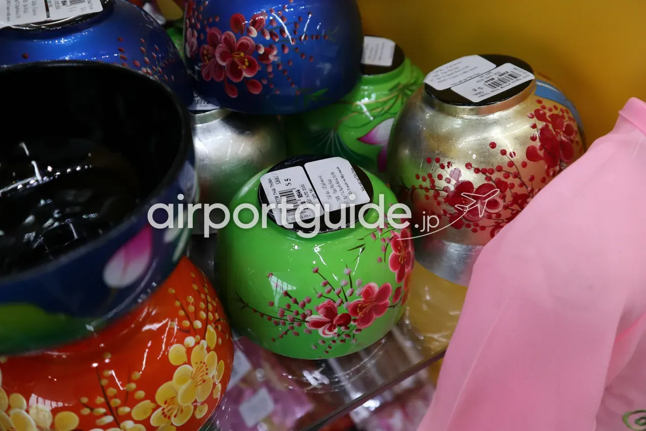 空港土産店の棚に並ぶベトナム漆器のクローズアップ。青・緑・金・オレンジの漆塗りの鉢に、赤やピンクの梅・桜の花柄が手描きされ、「$5」の値札が付く。