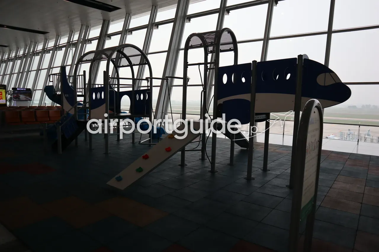 ノイバイ空港制限エリアの子ども遊び場。青い飛行機型の遊具とすべり台が、駐機場を望む床から天井までの大きなガラス窓の逆光の中に立つ。奥に霧のかかった駐機場。