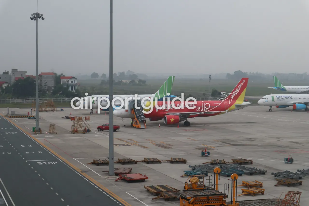ノイバイ空港の駐機場を窓から俯瞰。バンブーエアウェイズのA320、赤・黄のベトジェットエア（VietjetAir.com「Giá rẻ hơn」）のA320、もう1機のバンブー機が並び、手前に手荷物ドーリーや地上支援機材、「STOP」の誘導路標示。