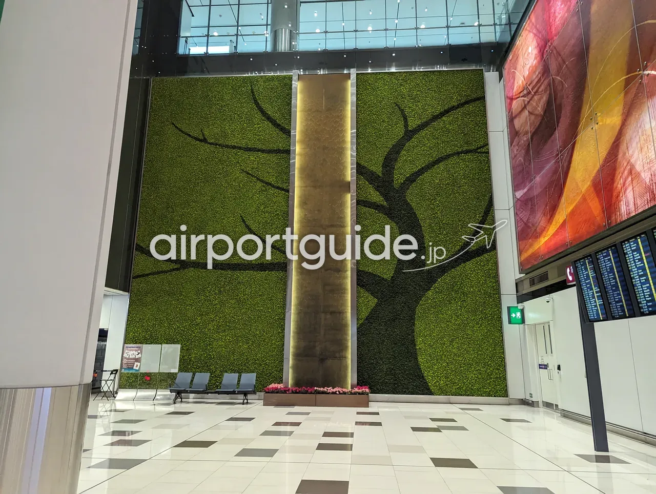 香港国際空港の館内アート。天井まで届く大きな緑化壁に樹木のシルエットが描かれ、中央に金色の滝状オブジェ、足元に花壇とベンチ。右手に赤系の大型アートスクリーンとフライト情報の電光掲示。市松模様の石床。