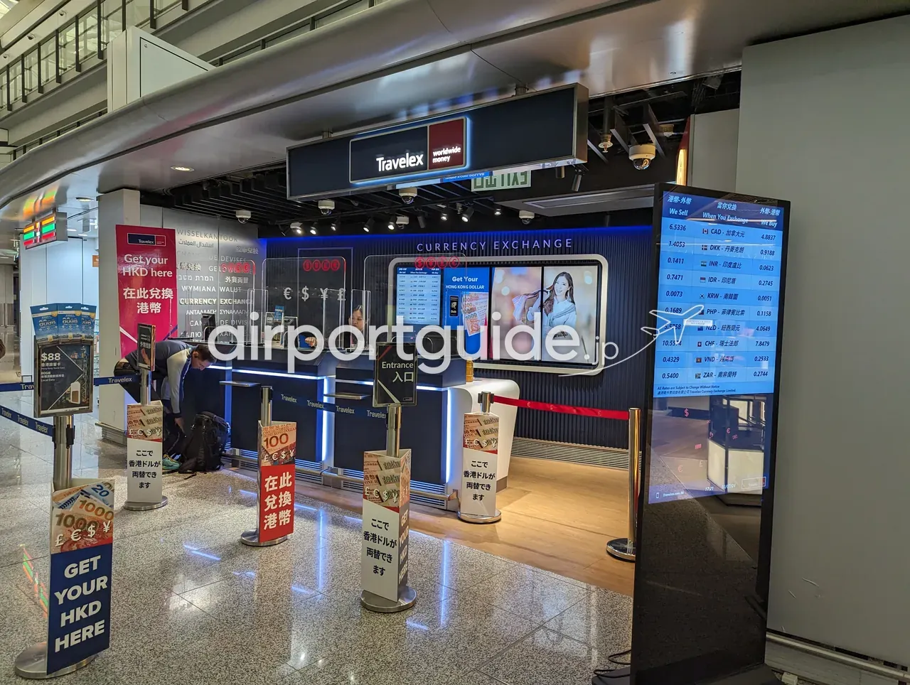 香港国際空港のTravelex両替所を別アングルから。各国通貨の売買レート電光板と『GET YOUR HKD HERE』の案内が見える。
