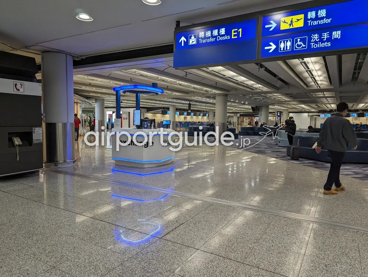 香港国際空港の乗り継ぎエリアにある『exchange 兌換』の両替キオスク。頭上に『Transfer Desks E1 / Transfer / Toilets』の案内サイン。
