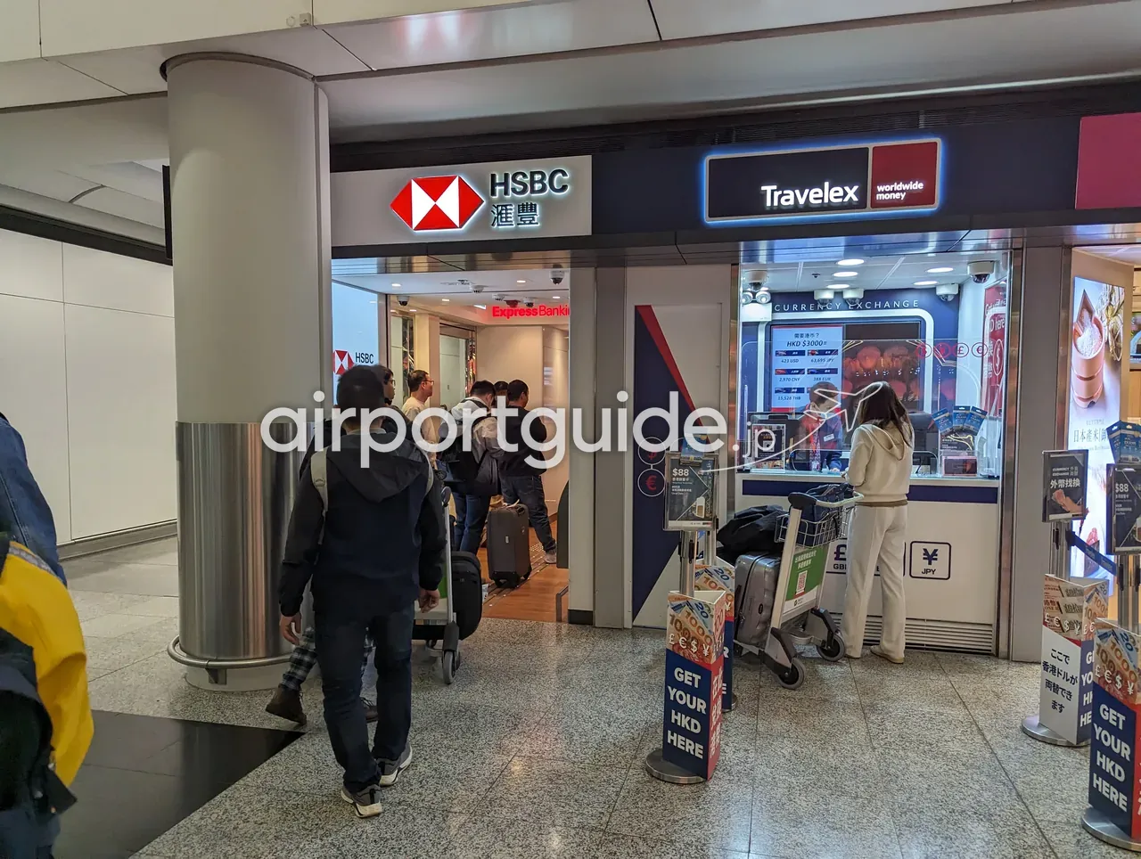 香港国際空港の通路。左にHSBC（滙豐）の支店『Express Banking』、右にTravelexの両替所『CURRENCY EXCHANGE』が並び、『GET YOUR HKD HERE』の案内。荷物カートやスーツケースを持つ利用客が行き交う。