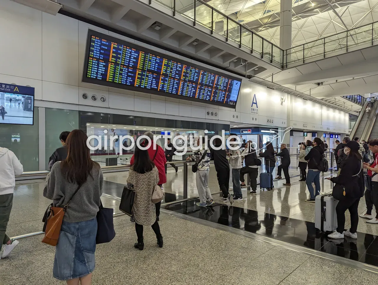 香港国際空港・接機大堂（Arrivals Hall A）の大型到着便案内板の前で、出迎えの人々が手すり沿いに並ぶ。天井は三角格子の大屋根、右手にエスカレーター、『大堂出口 Hall Exit A』の表示。