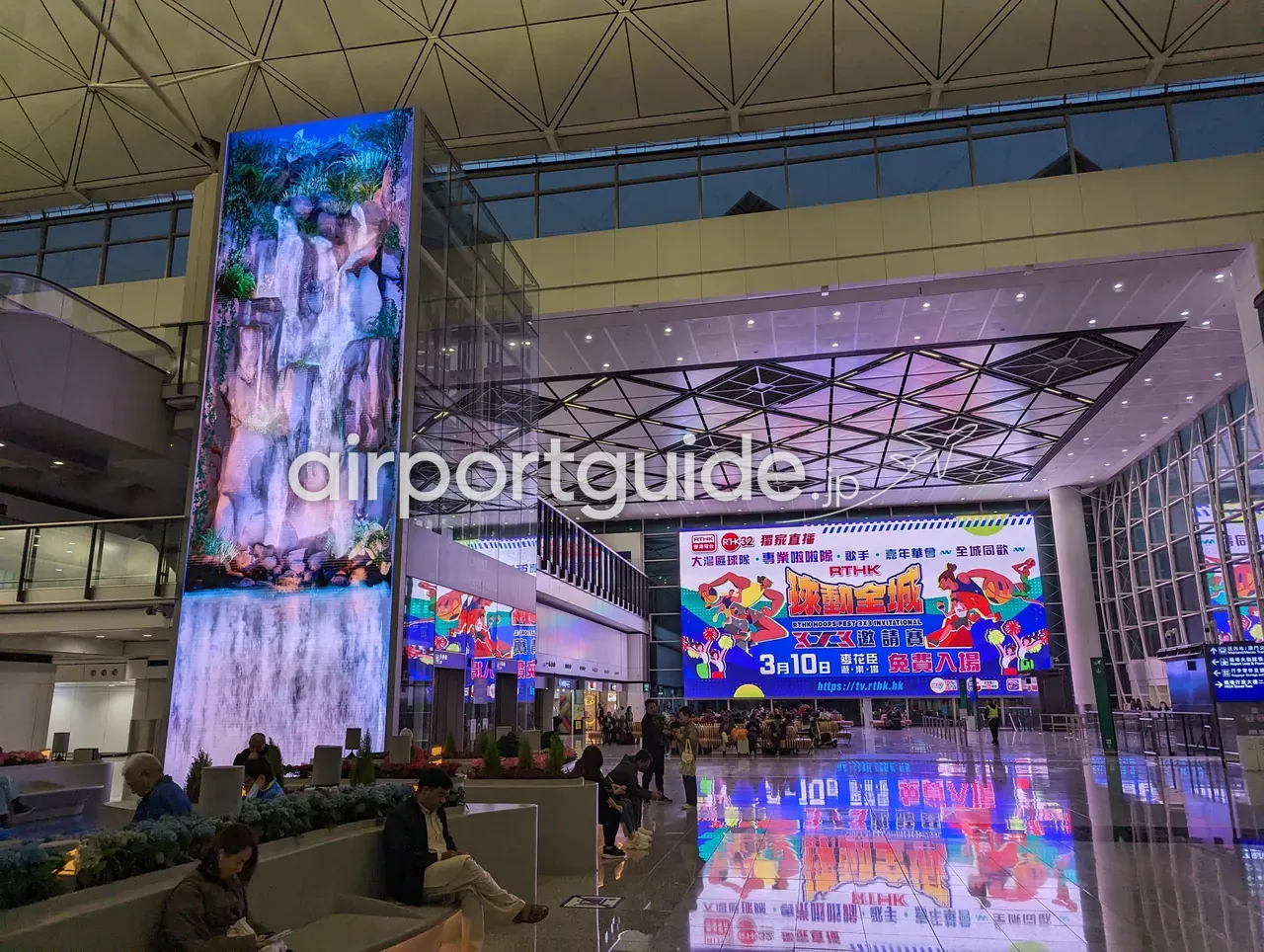 香港国際空港の縦長のデジタルアート塔。滝の映像を映す高いLEDの柱と、右手に大型広告LED、円形ソファでくつろぐ人々、天井は三角格子のガラス、床に映り込み。