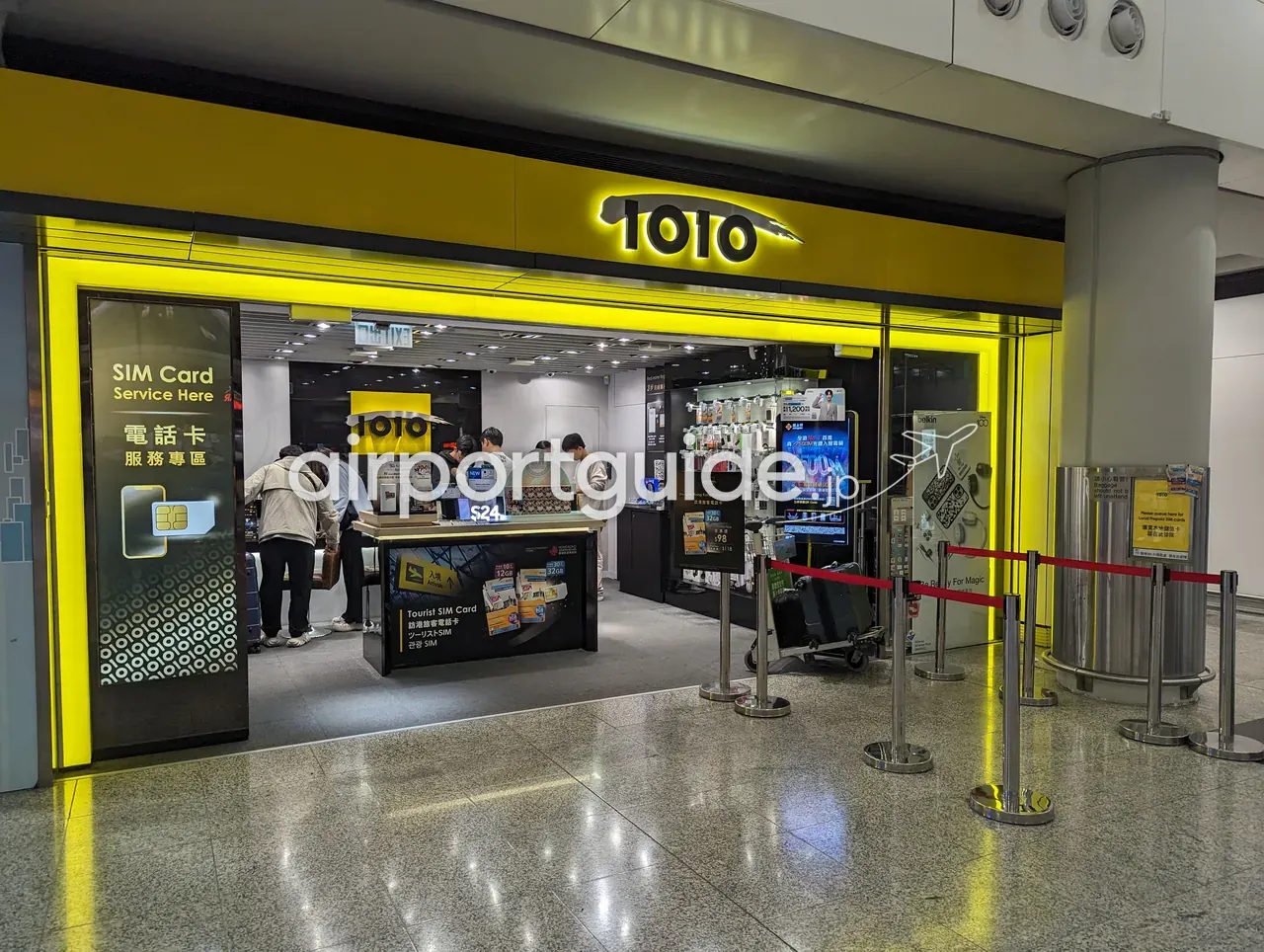 香港国際空港の携帯・SIMカード店『1O1O』。黄色い店構えに『SIM Card Service Here 電話卡服務專區』、店頭カウンターに『Tourist SIM Card（ツーリストSIM）』のパッケージ、店内の客とスタッフ。