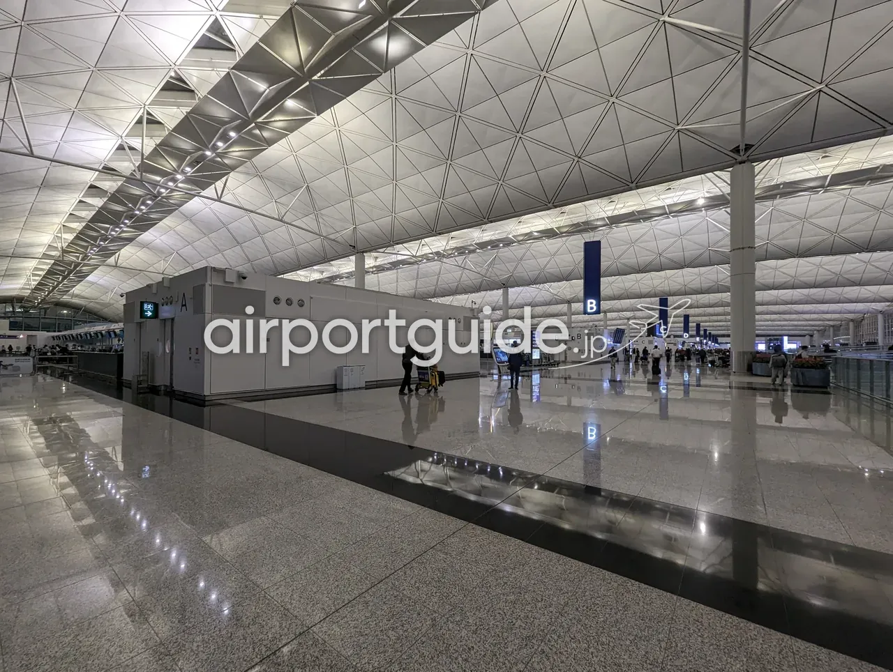 香港国際空港・ターミナル1の出発チェックインホール。三角格子を組んだ波打つ巨大な大屋根の下、青い柱状のアイル標識A・B・C・Dが並び、奥にチェックインカウンター列と荷物カートを押す旅客。磨かれた御影石の床。