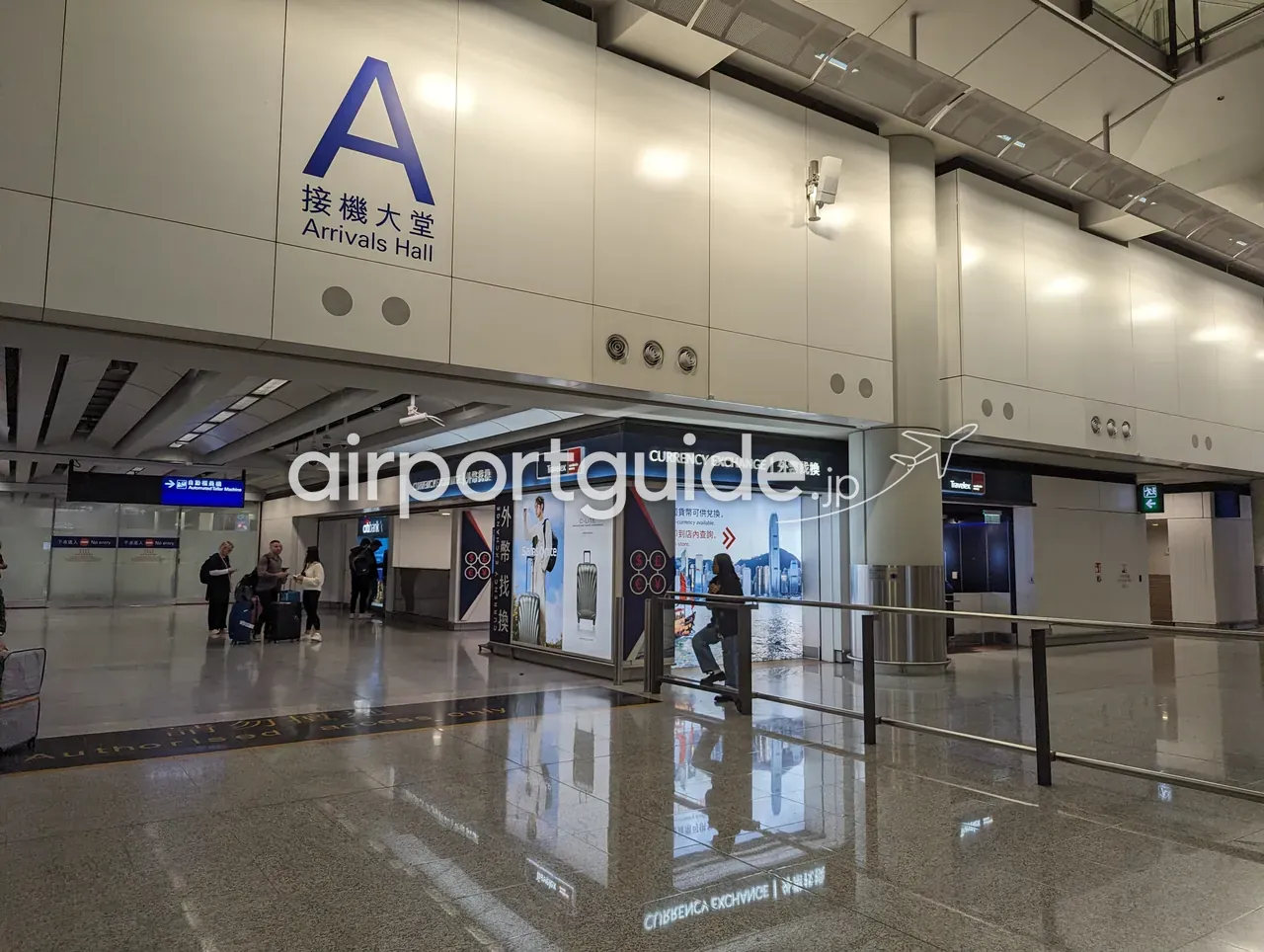 香港国際空港の到着ホール入口。大きな『A 接機大堂 Arrivals Hall』の表示の下に、Travelexの両替所『CURRENCY EXCHANGE 外幣找換』のカウンターがある。手前をスーツケースの旅客が歩く。