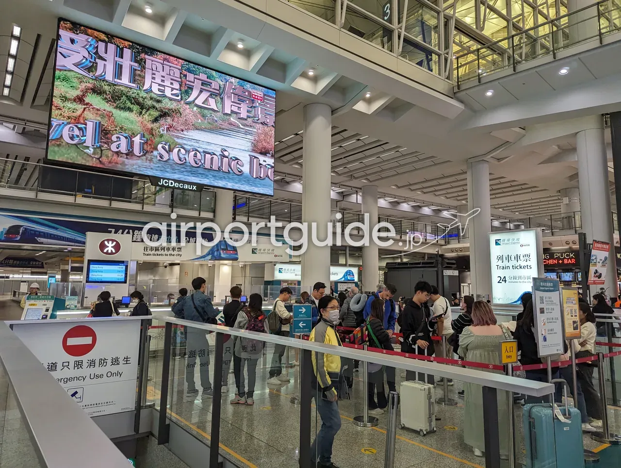 香港国際空港のエアポートエクスプレス切符売り場。MTRロゴと『機場快綫 Airport Express』『往市區列車車票 Train tickets to the city』の窓口、右手に『24 minutes to the city（24分で市内へ）』の表示、頭上に大型映像広告、荷物を持つ長い列。