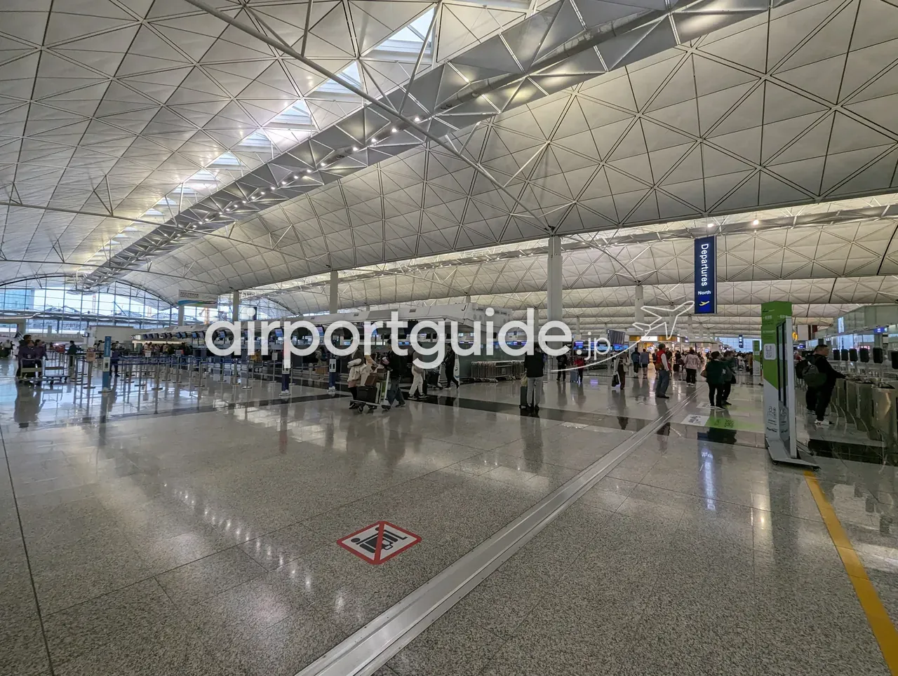 香港国際空港・ターミナル1の出発チェックインホール。三角格子の巨大な大屋根の下、青い吊りサイン『Departures North』、ずらりと並ぶチェックインカウンター島、荷物カートの旅客、右手にセルフチェックイン機。