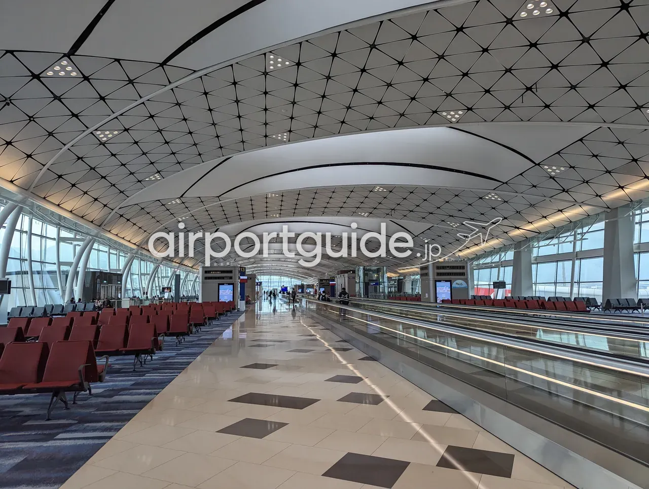 香港国際空港・制限エリアの搭乗ゲート付近のコンコース。曲面を描く格子天井の下、両脇に赤いゲート待合座席が並び、中央に動く歩道（トラベレーター）。ゲート213付近、ほぼ無人の落ち着いた空間。