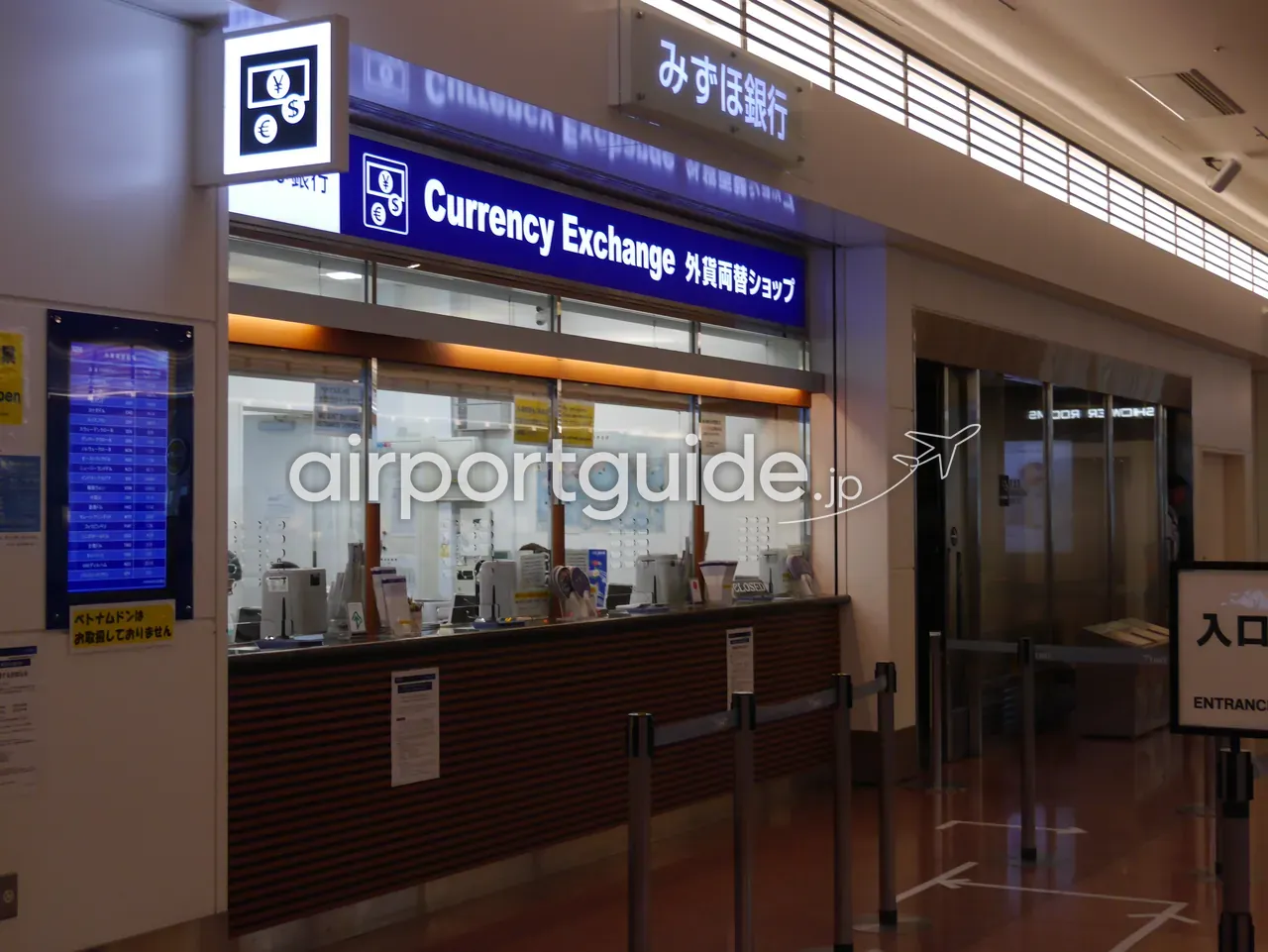 空港のみずほ銀行「Currency Exchange 外貨両替ショップ」の窓口。左手に円貨から外貨への為替レート板、奥の壁に世界地図が掲げられている。