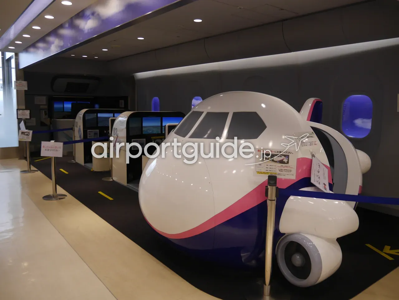羽田空港の体験コーナーに並ぶ、飛行機の機首をかたどったフライトシミュレーターの筐体。雲を描いた天井の下、順番待ちのレーン。