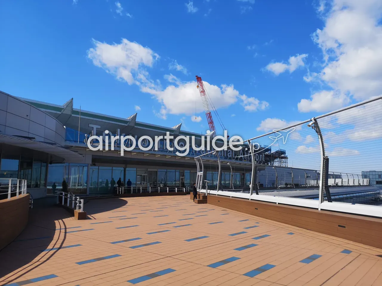 羽田空港の屋上展望デッキ。ウッドデッキの床と金網フェンス、建物壁面に『Tokyo International Airport』の文字、快晴の青空。