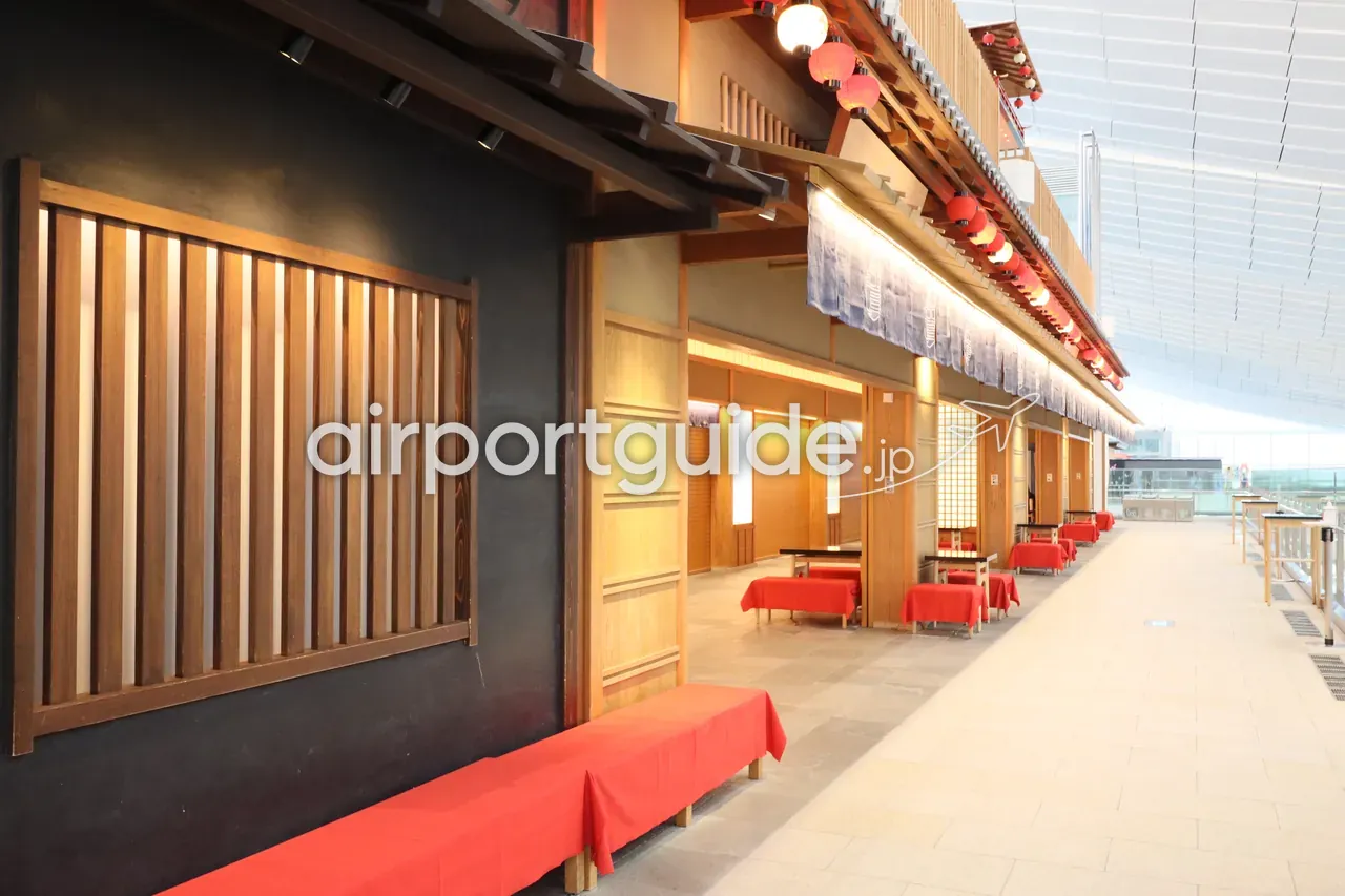 羽田空港第3ターミナル『江戸小路』の飲食エリア。黒壁と木格子の江戸町屋風のファサードに、赤い毛氈を掛けた縁台と赤提灯、藍染めの暖簾が並ぶ。
