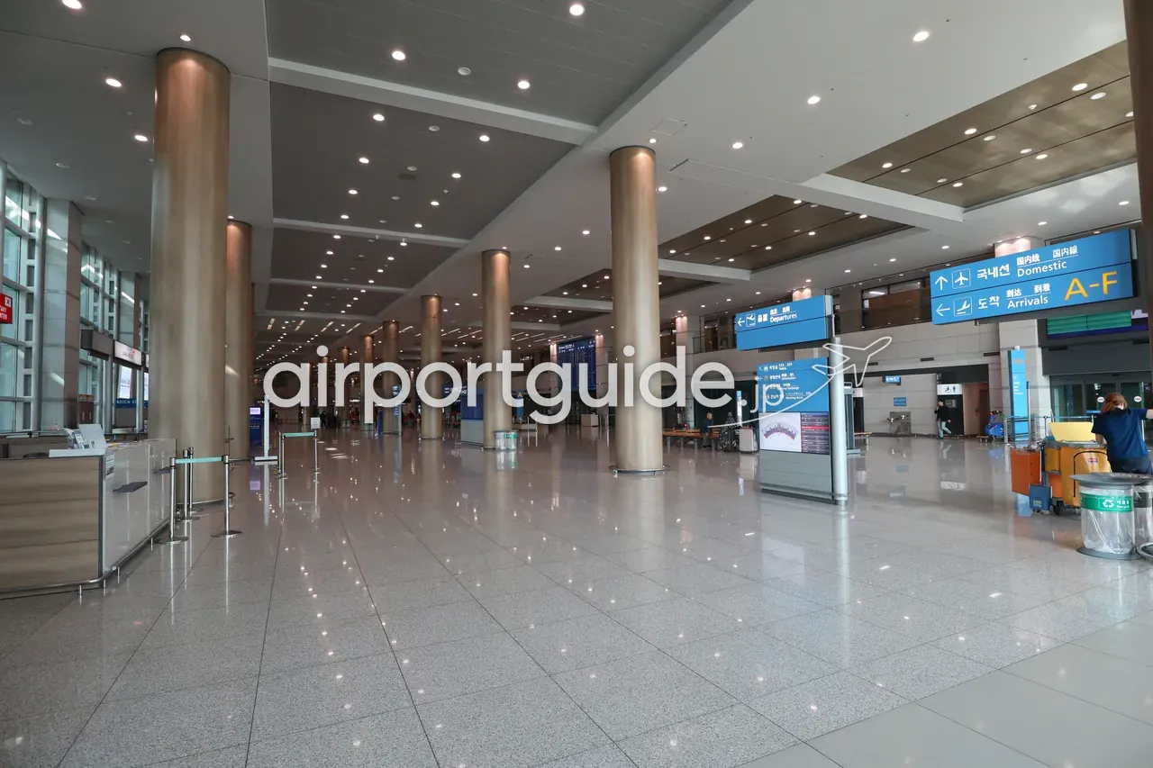 仁川空港の連絡ホール。木目調の丸い柱が並ぶ大空間に、「国内線 Domestic」「到着 Arrivals A-F」などの青い吊り下げ案内サイン（韓・日・英・中）が下がる。