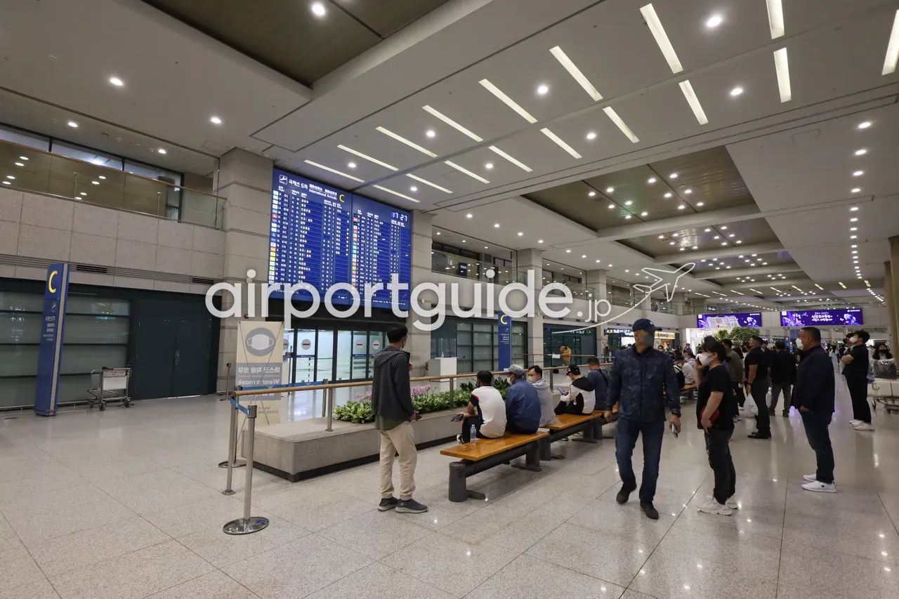 仁川空港の到着ロビー（出口C付近）。大型の到着便案内板の下で、ベンチに座る人やスマホを見る人が到着客を待つ。