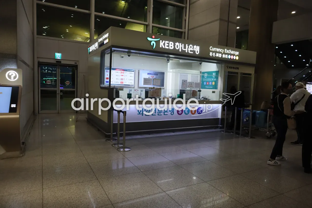 夜の仁川空港、KEBハナ銀行の両替ブース。右手にスーツケースの旅行者が待ち、左にセルフの案内キオスクが立つ。