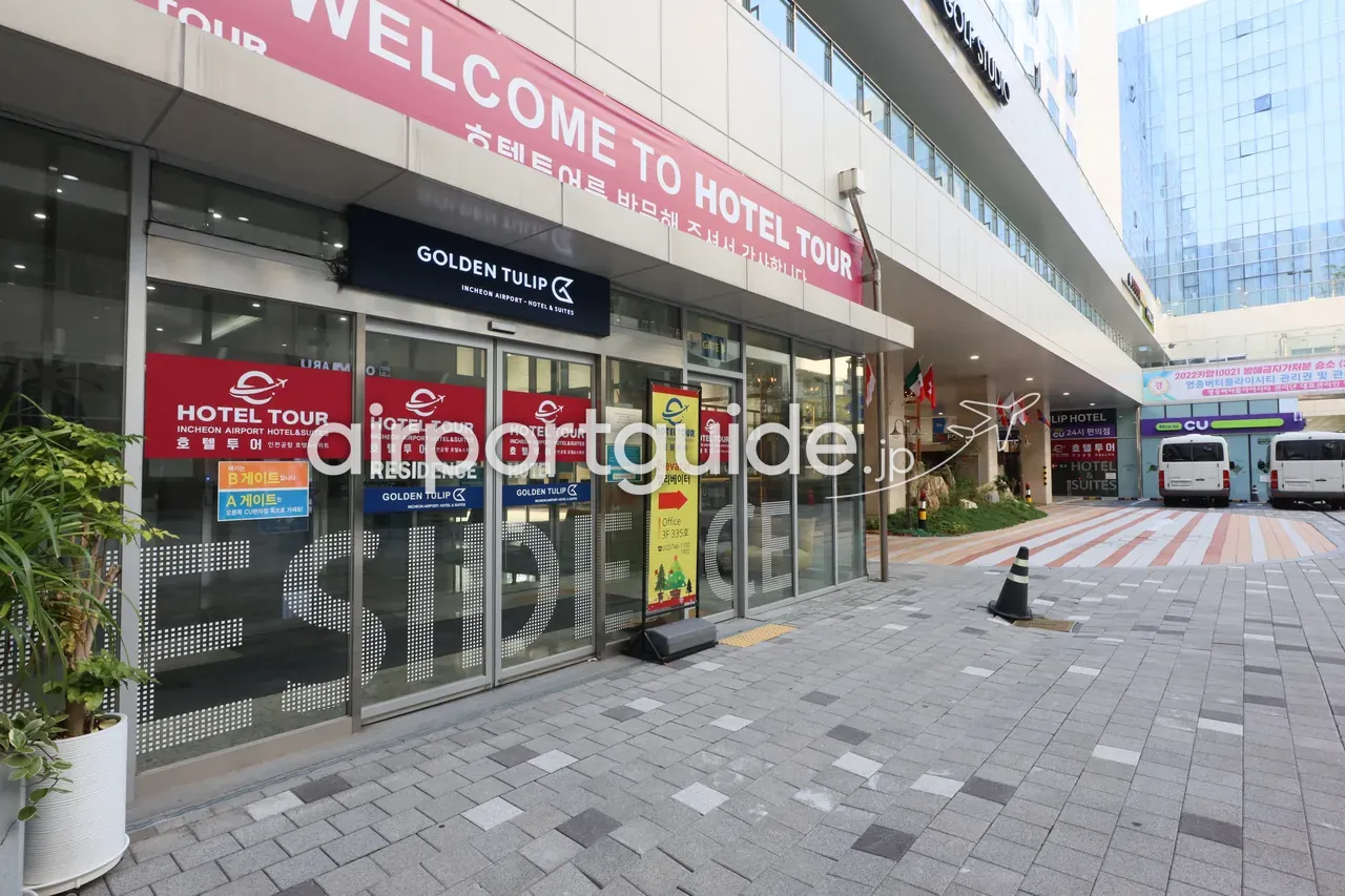 ゴールデンチューリップ仁川空港ホテルの外観・入口。「GOLDEN TULIP INCHEON AIRPORT · HOTEL & SUITES」の黒看板、右手にCUコンビニと白いシャトルバン、各国の国旗。