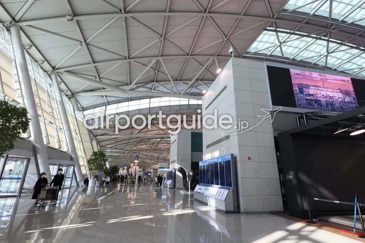 仁川空港第1ターミナルの出発ホール。曲面トラスの大屋根の下、フライト情報板と大型広告スクリーン、Gゾーンのチェックインエリアが広がる。