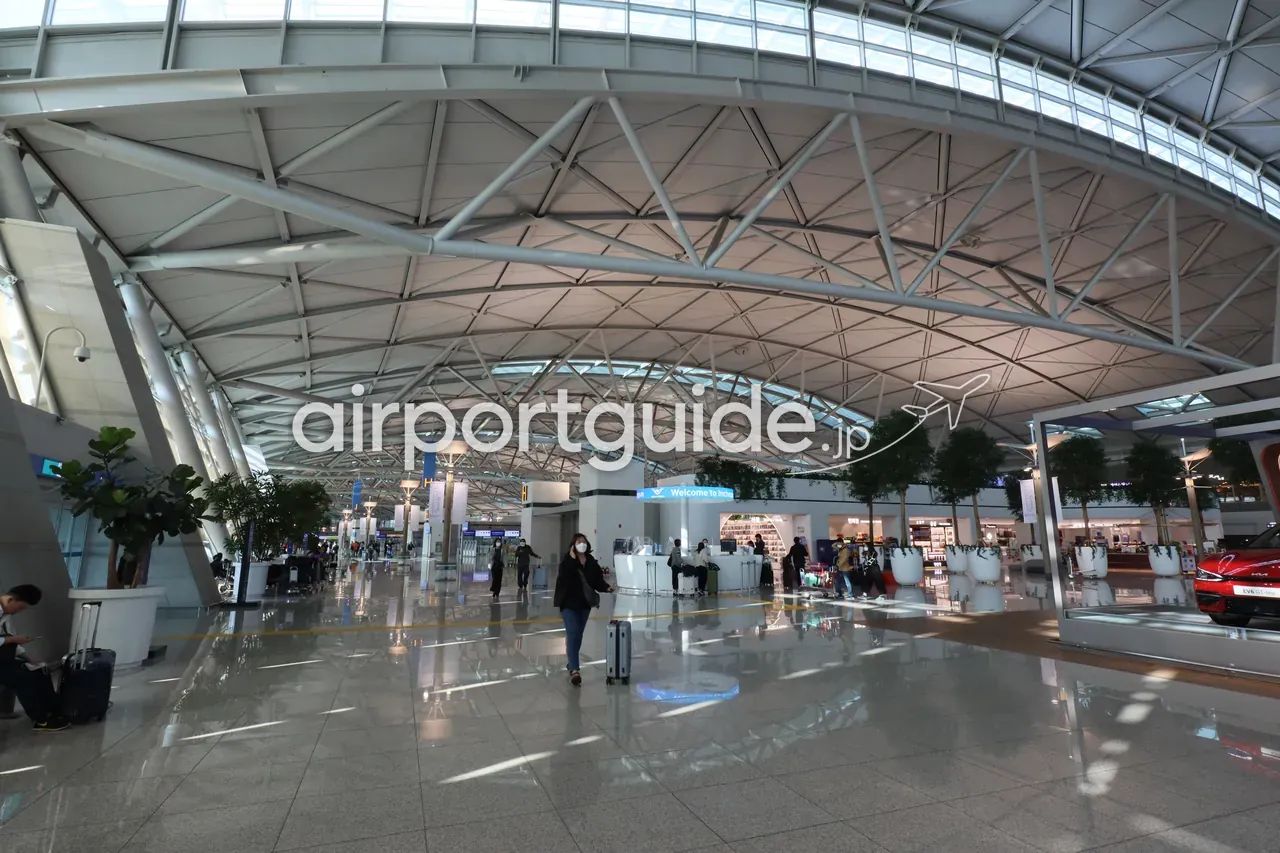 仁川空港第1ターミナルの出発ホール。曲面トラスの大屋根の下、「Welcome to Incheon」のサインと免税店、右手に赤いEV車の展示が並ぶ広い空間。