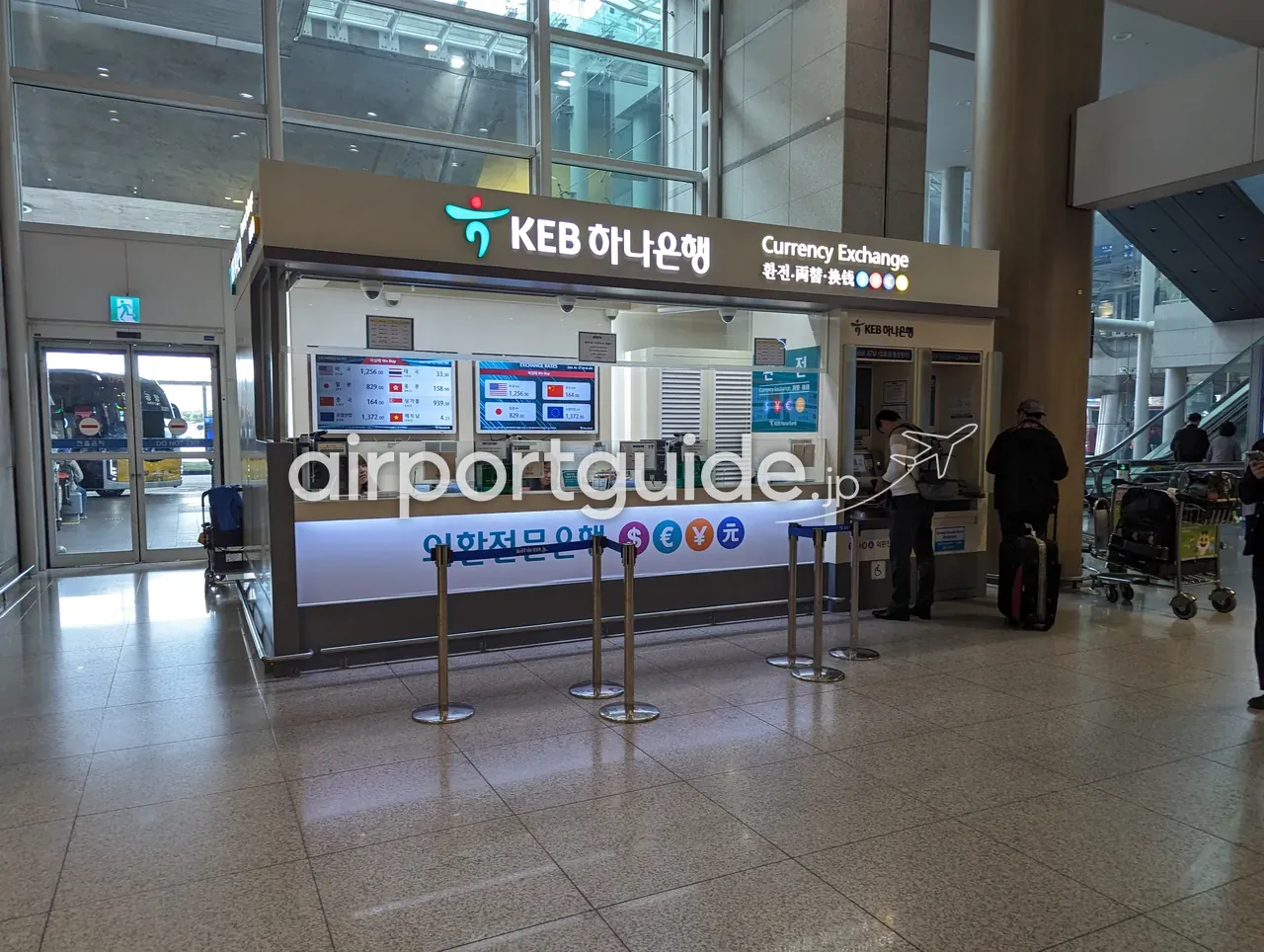 仁川空港第2ターミナル、KEBハナ銀行の到着エリア両替カウンター。為替レートの電光ボードとGlobal ATMが並び、スーツケースの旅客が手続きしている。