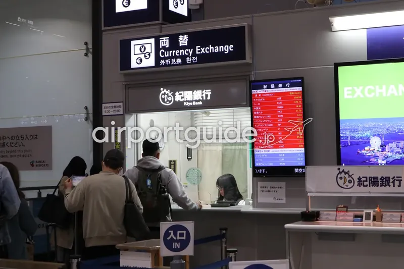 関西空港の紀陽銀行（Kiyo Bank）両替所。「両替 Currency Exchange」看板の下に為替レートモニターが並び、旅客が窓口に列を作っている。
