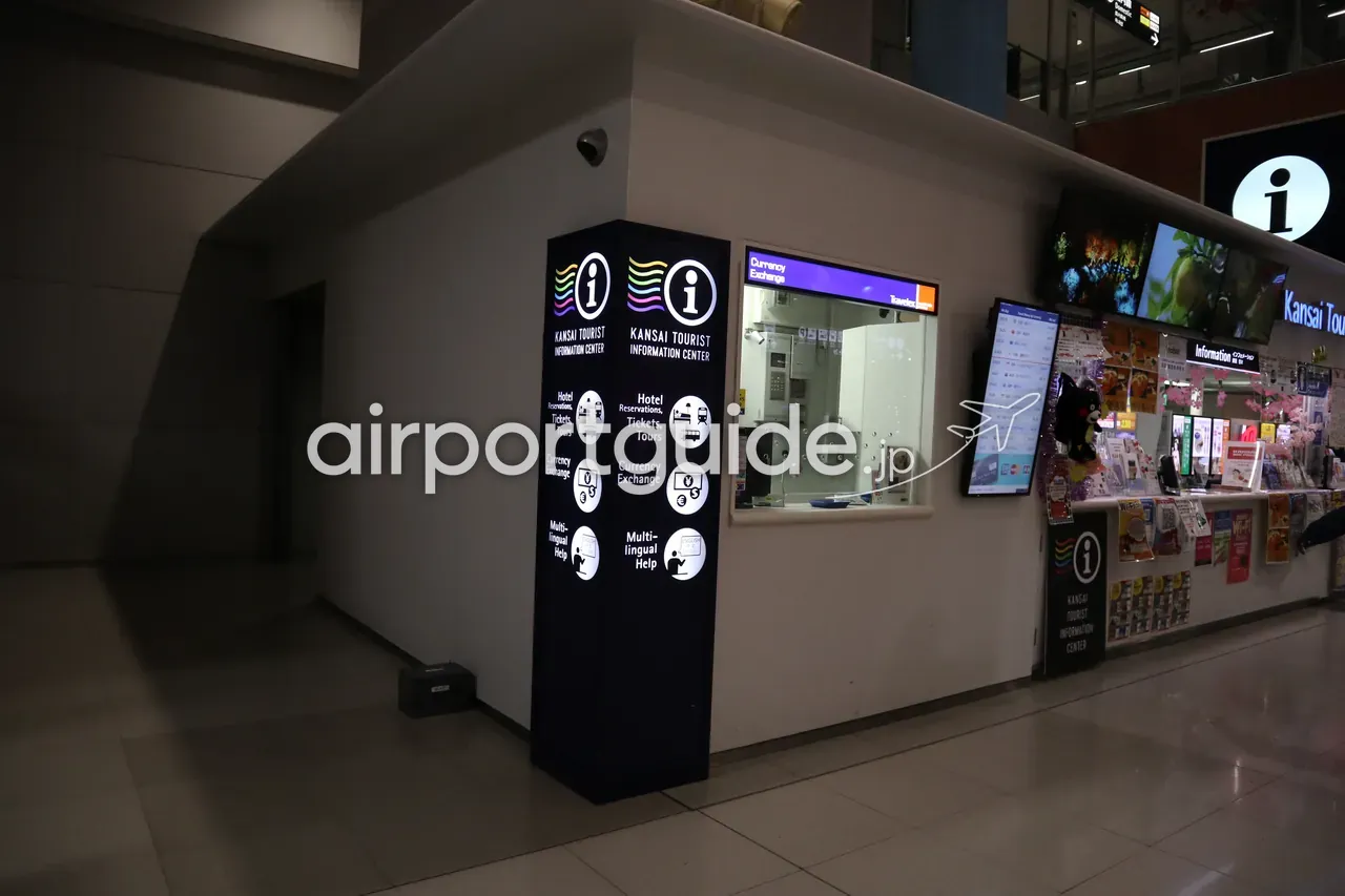 関西空港の関西ツーリストインフォメーションセンター。「i」マークのピラーサインに、ホテル予約・両替・多言語対応を示すアイコンと、パンフレット棚のあるカウンター。