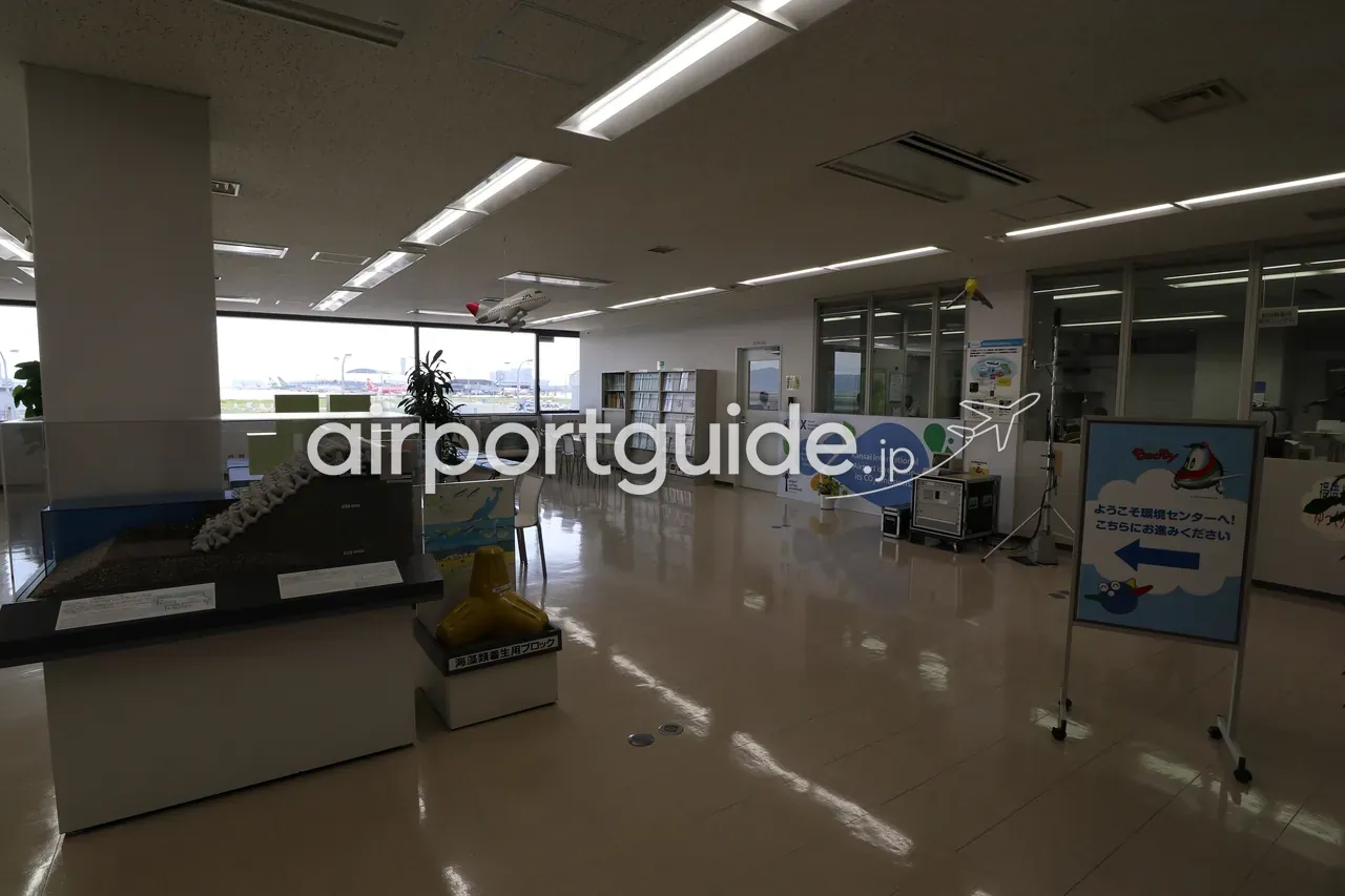 関西空港 環境センターの展示スペース。空港のCO2削減の展示パネルや海中鉄筋の模型、右手のガラス越しに職員の執務室、窓の外に駐機中の機体。