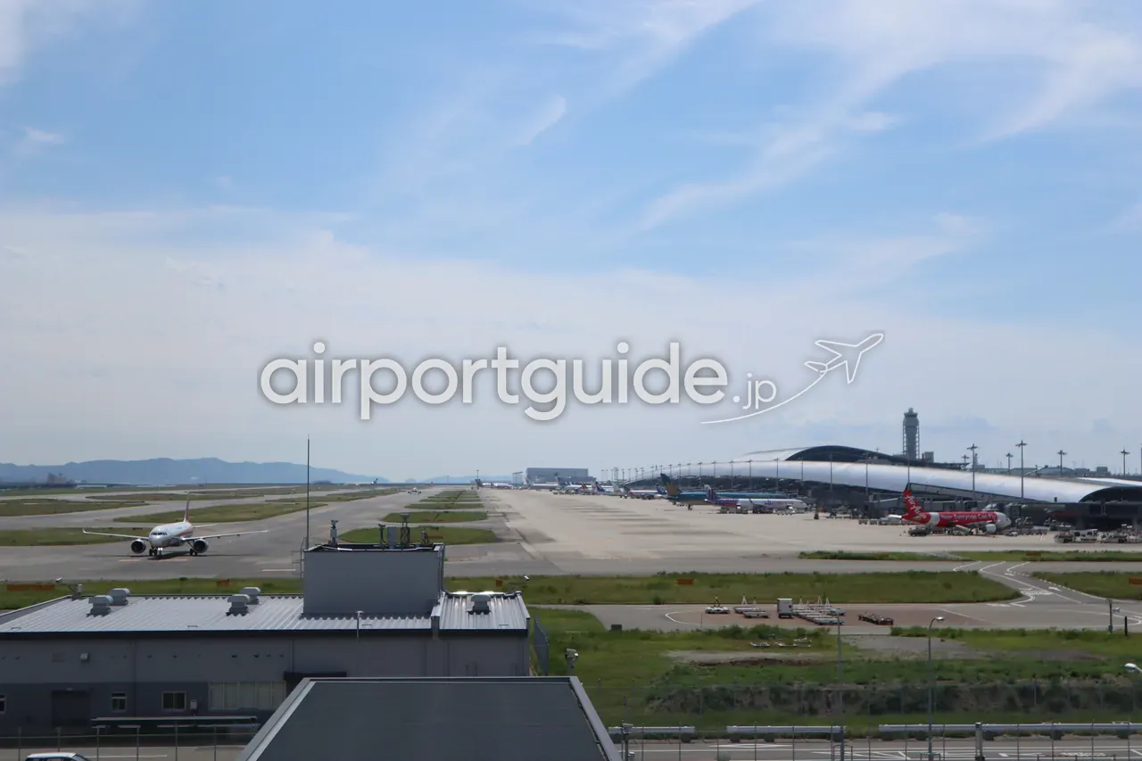 スカイデッキから望む関西空港のエプロンと第1ターミナル。駐機中の機体、誘導路を走る旅客機、右奥に管制塔と波形の大屋根。