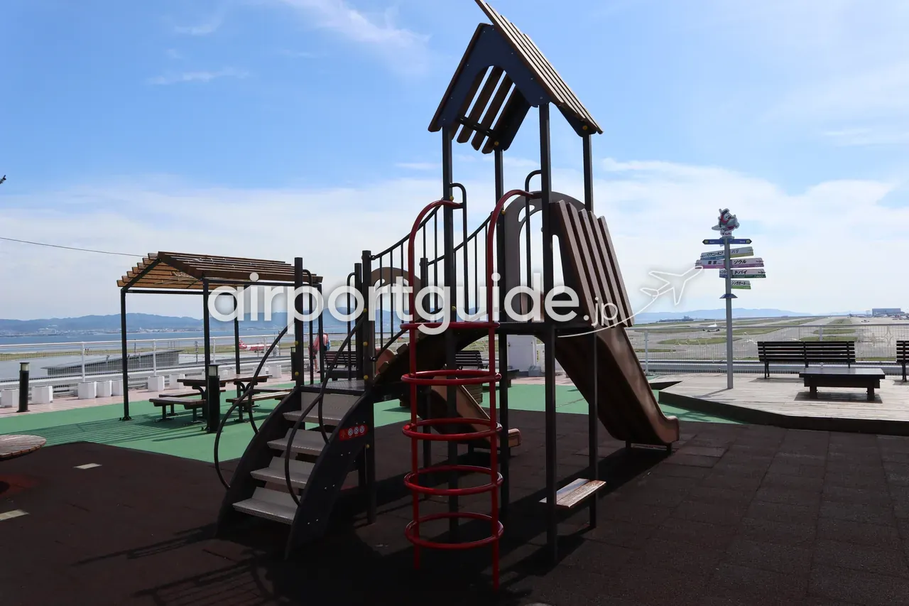 スカイビュー屋上の子ども向けプレイグラウンド。木製の複合遊具と滑り台、奥に滑走路と海、右手に各都市への方面を示す標識。
