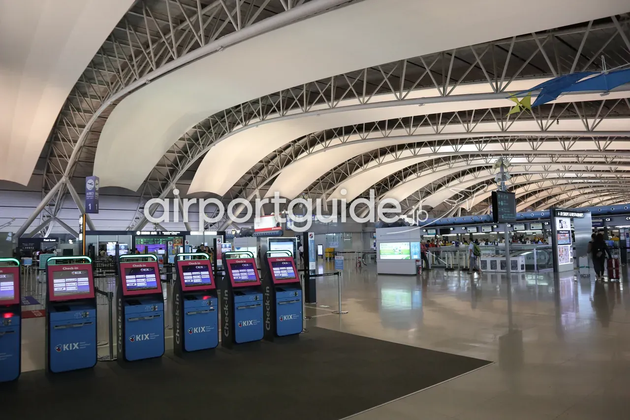関西空港 第1ターミナルの出発ロビー。手前に「KIX Check-in」自動チェックイン機の列、うねる波形の大屋根の下にカウンターと行き交う人。