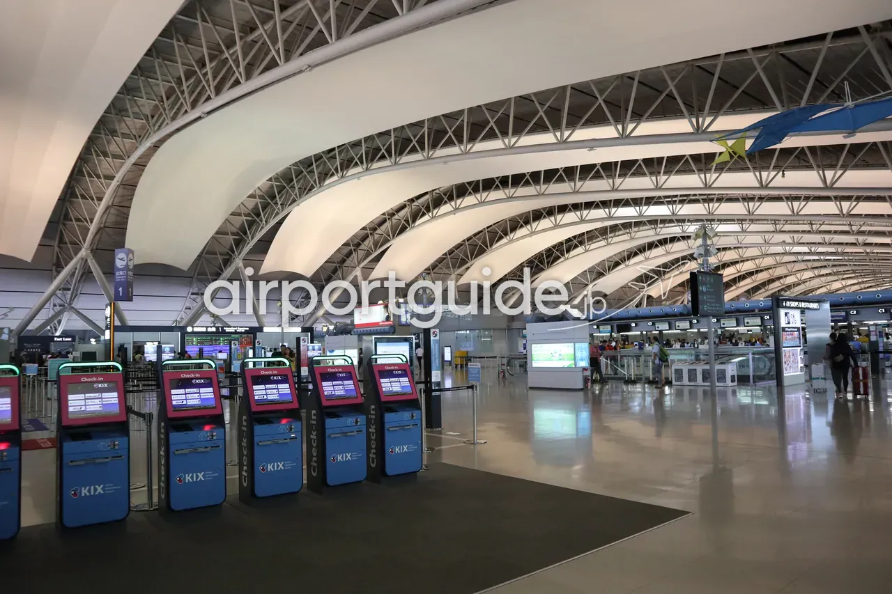関西空港第1ターミナルの出発ロビー。うねる波形の大屋根の下、手前に青い「KIX Check-in」自動チェックイン機の列が並び、奥にチェックインカウンターと旅客。
