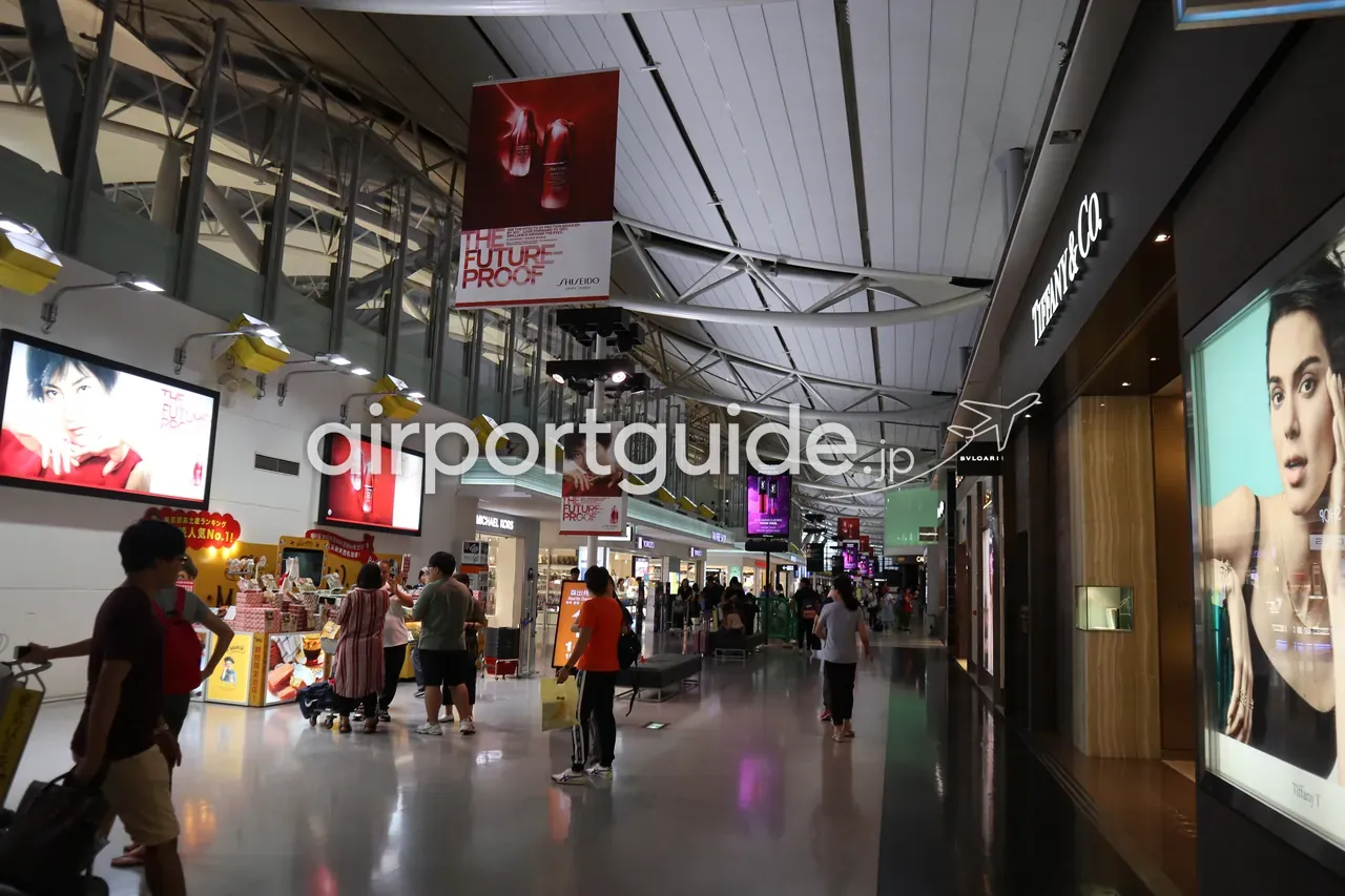 関西空港の制限エリア内の免税店街。資生堂の広告や、ティファニー・ブルガリなどのブランドブティックが並び、旅客が行き交う波形屋根の通路。