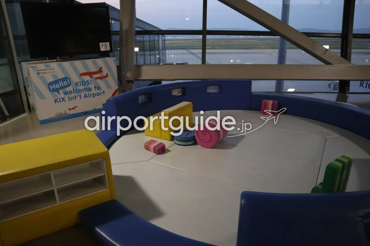 関西空港の搭乗待合エリアにある円形のキッズプレイエリア。青いクッションの座面と「Hello! KIDS! welcome to KIX Int'l Airport」のパネル、窓の外は夕暮れの駐機場。