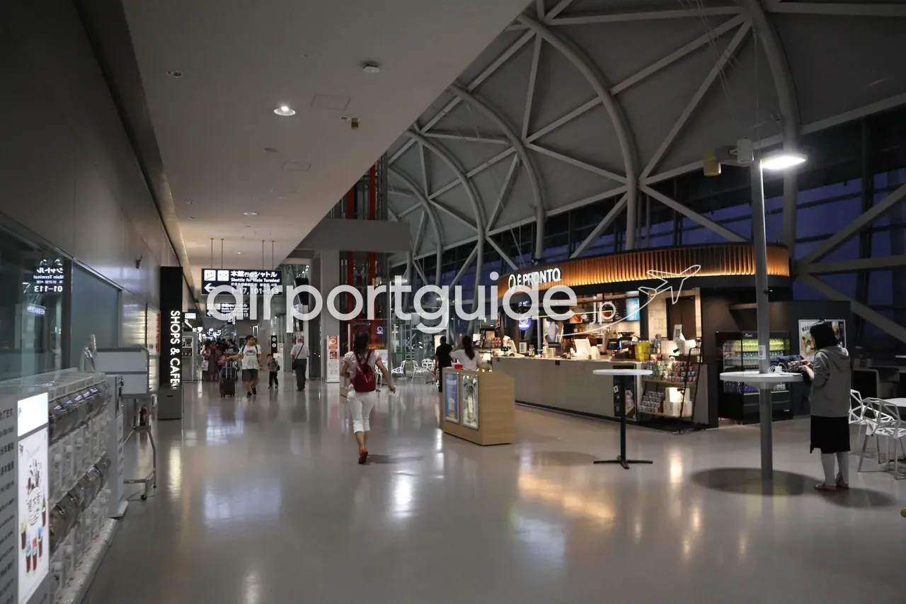 関西空港のコンコース。右手にカフェ「PRONTO」、天井は連絡橋風のトラス構造で、左に「SHOPS & CAFE」の案内、数人が歩く。