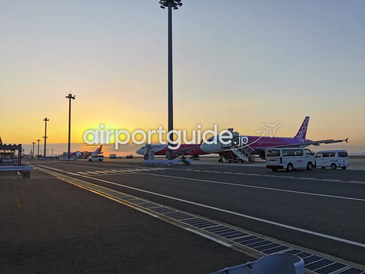 夕日が沈む関西空港のエプロン。ピンク色のPeach（ピーチ）のエアバスA320型機とタラップ、ランプバスが並び、奥に大阪湾。