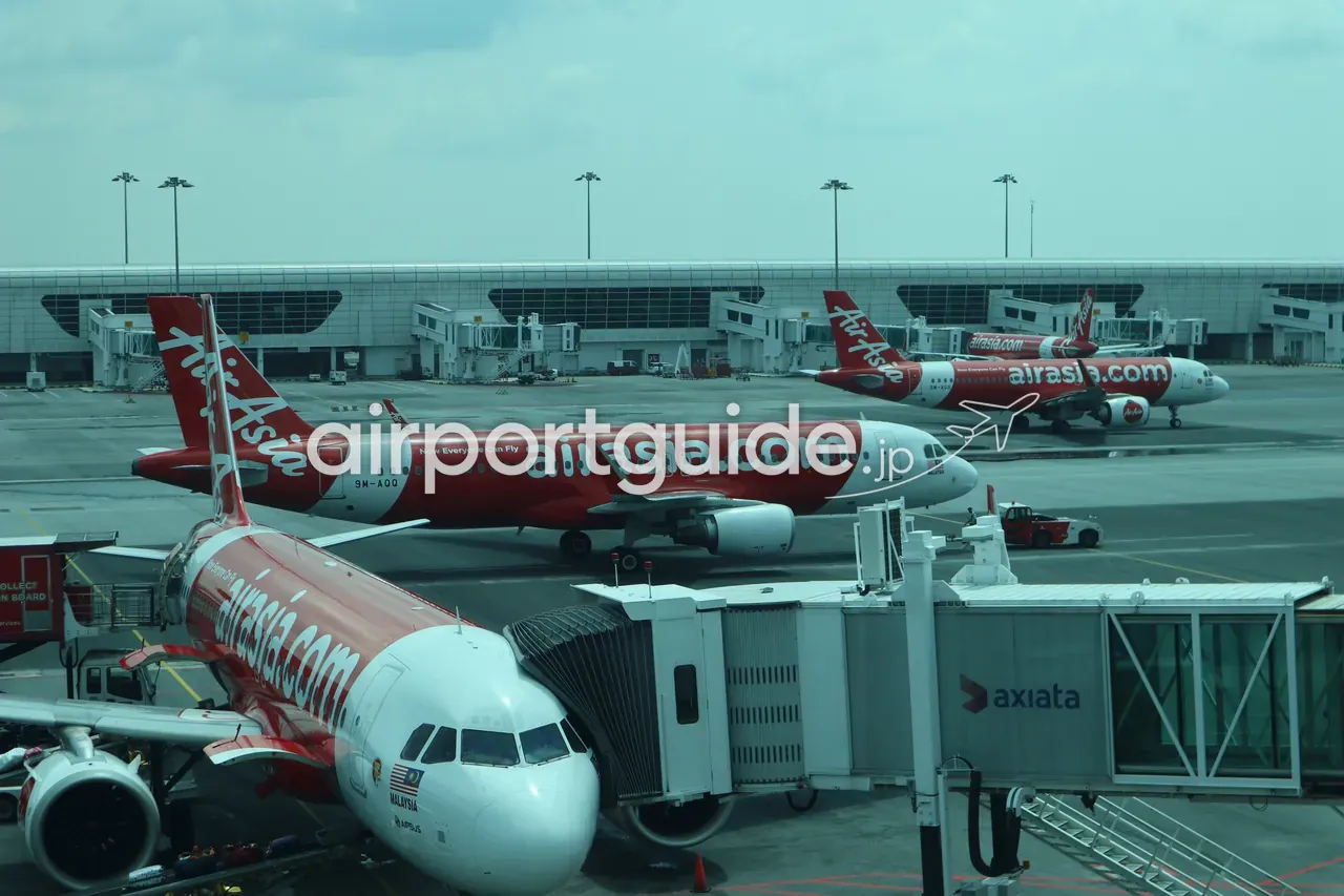 ターミナル2のエプロンを昼間に望む眺め。「airasia.com / Now Everyone Can Fly」塗装のエアバスA320が複数、ボーディングブリッジに接続して駐機し、地上作業車が動く。