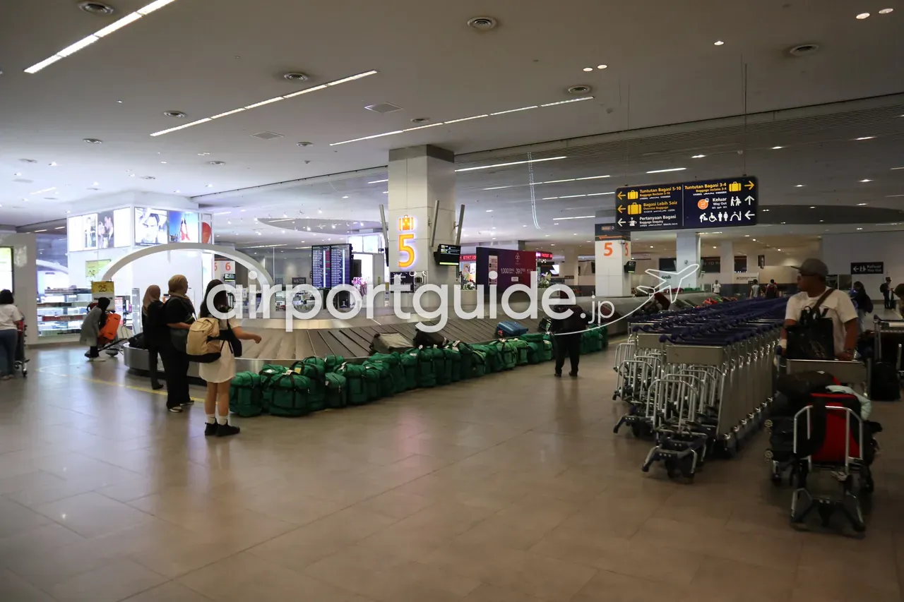 KLIAの手荷物受取エリア。ベルト5番のターンテーブルに緑のダッフルバッグが並び、「Tuntutan Bagasi / Baggage Reclaim」「Keluar / Way Out」の案内サインと、整列した手荷物カート。