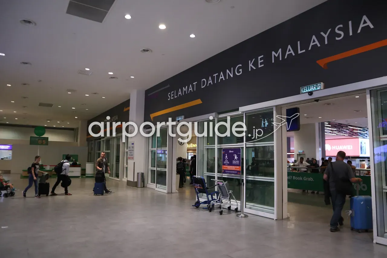 KLIAターミナル2の到着出口。壁面に大きく「SELAMAT DATANG KE MALAYSIA / WELCOME TO MALAYSIA」の歓迎サイン、ガラス自動ドアを抜ける旅客、右手にOldTownやHuaweiの店、Grabの案内。