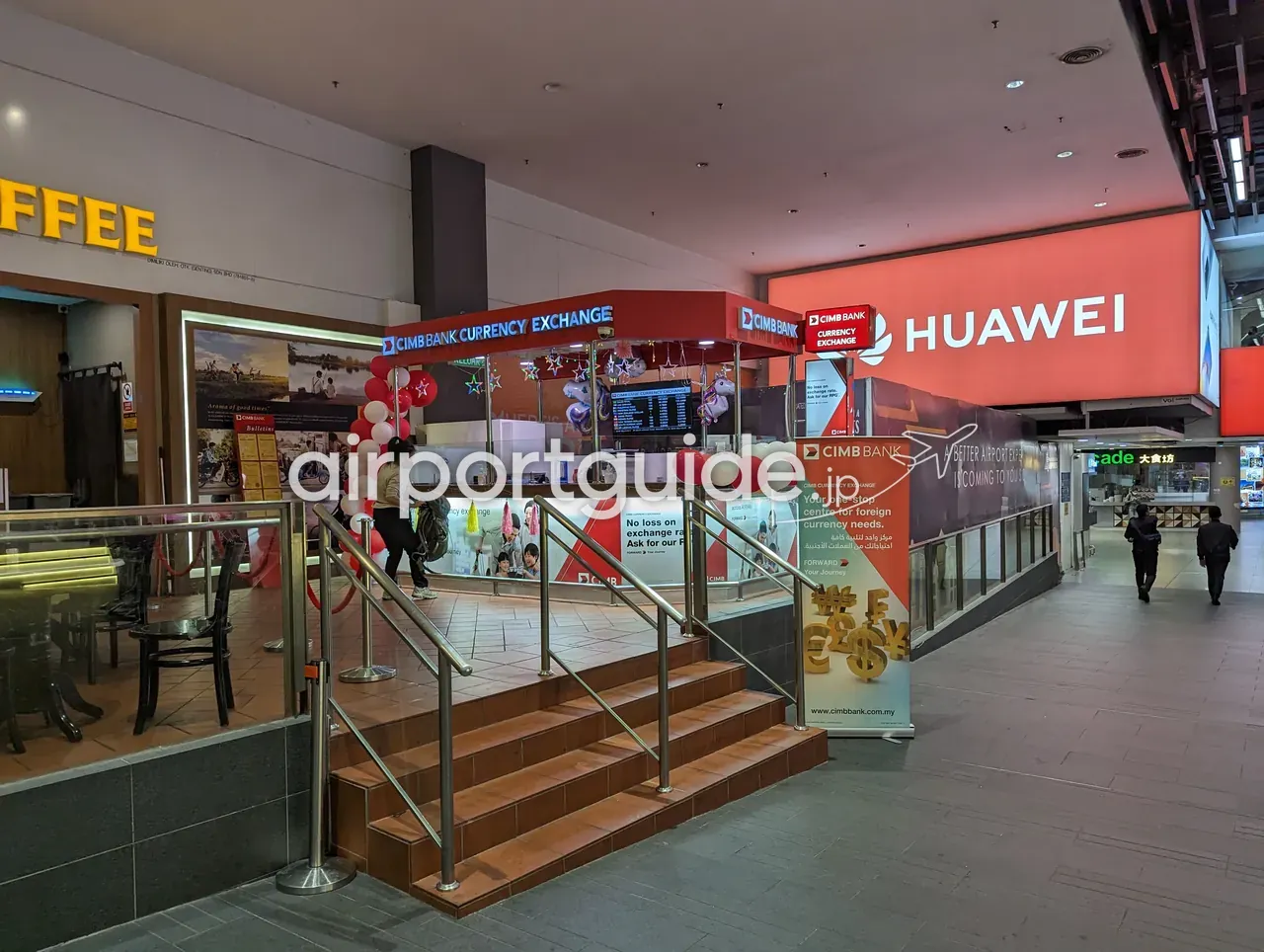 gateway@klia2モールのコンコースに立つCIMB Bank Currency Exchangeのブース。赤い庇に「CIMB BANK CURRENCY EXCHANGE」、赤白のバルーン装飾とデジタルのレートボード。手前に「CIMB BANK／Your one-stop centre for foreign currency needs」の縦看板、リュックを背負った旅行者が数段の階段を上って窓口へ向かう。奥に大きな赤いHUAWEIの広告ビジョン。