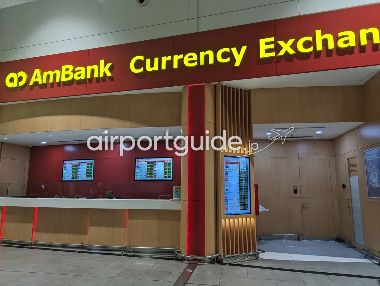 KLIA2のAmBank Currency Exchangeカウンター。赤地に黄色の大きな「AmBank Currency Exchange」看板、木目とワインレッドの内装。窓口上に2枚の為替レートモニタ、右手前の柱に多通貨のBank Buy/Bank Sellを縦一覧にした電光レート表示。