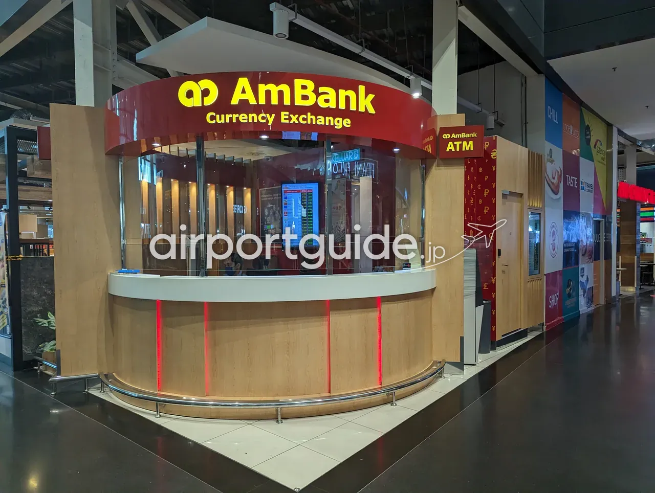 KLIA2・gateway@klia2の丸型のAmBank Currency Exchangeブース。赤い曲面の看板に黄色で「AmBank Currency Exchange」、木目のカウンターに赤いLEDのライン、窓口の中にデジタルレート板。右手に黄色い「AmBank ATM」の案内板とATMのコーナー、奥にモールの店舗サイン。