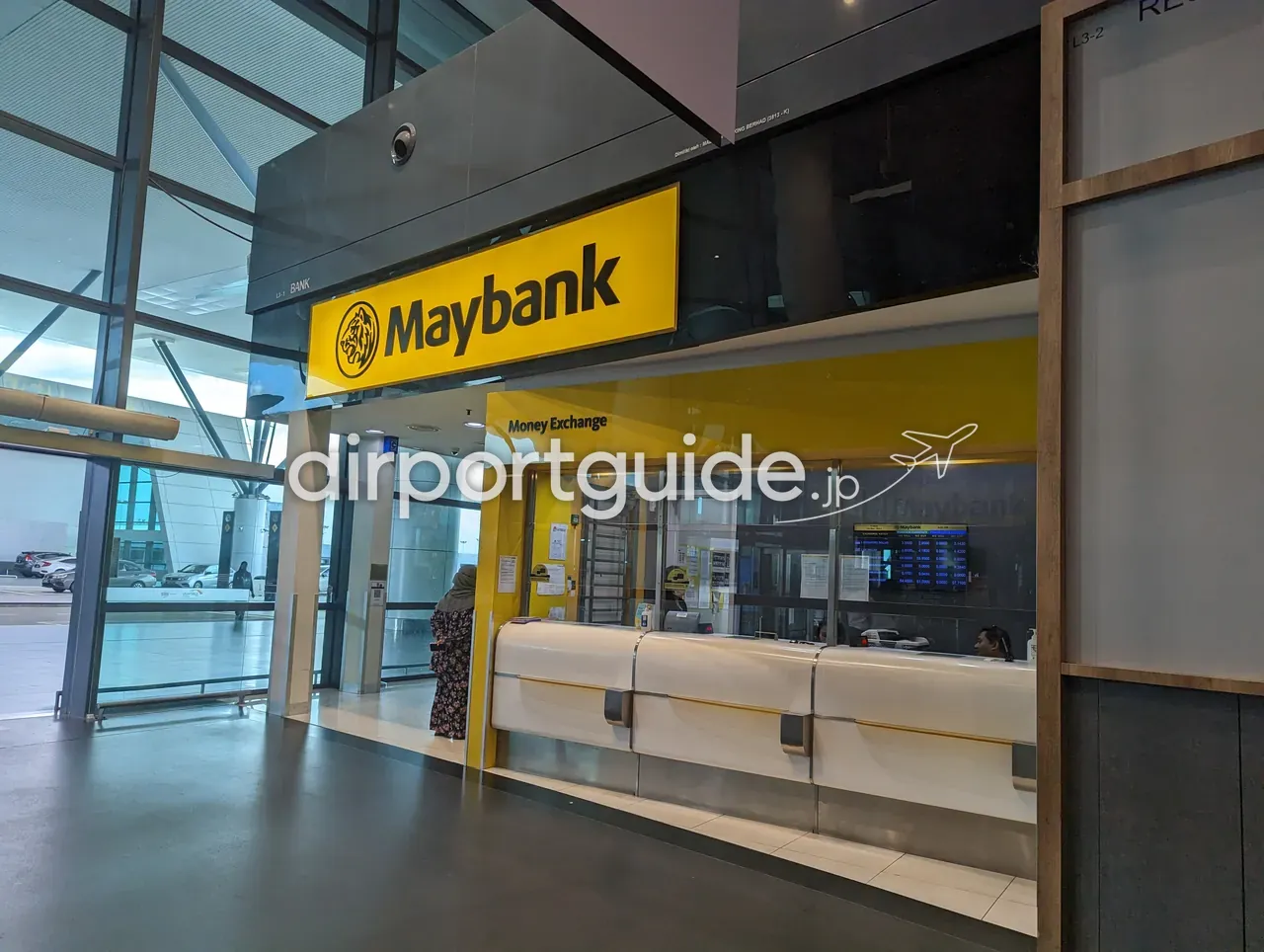 KLIA2のMaybank Money Exchangeカウンター。鮮やかな黄色の看板（トラの頭ロゴ）と「Money Exchange」のヘッダー、ガラス窓口の中にデジタルレートボード。左手は床から天井までのガラス窓で外の車寄せが見える。窓口前に柄物ワンピースの女性客が1人。
