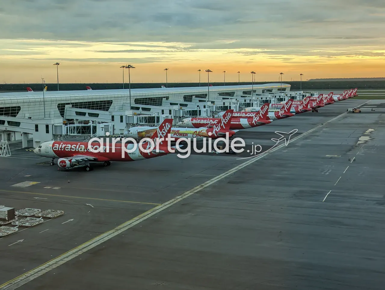 ターミナル2の展望窓から見た夕暮れのエプロン。ボーディングブリッジに沿って、赤い「airasia.com」塗装の機体が一列に並び、地平線がオレンジ色に染まる。